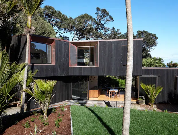 Exterior view of a contemporary elevated house surrounded by established coastal vegetation. The home has dark vertical timber cladding, projecting upper-level volumes, large glazed openings with warm timber surrounds, and a sheltered ground-level outdoor living area opening onto a landscaped lawn and garden.