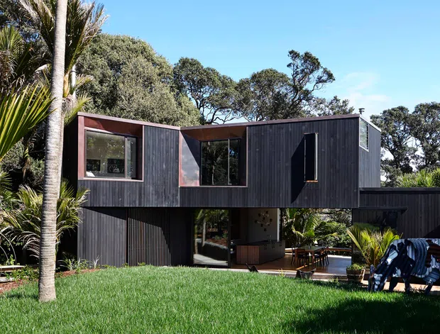 Exterior view of a contemporary two-storey house set within a lush, landscaped site. The building features dark vertical timber cladding, large windows, recessed openings, and a partially elevated upper level creating a covered outdoor area below. Tropical and native planting surrounds the lawn, with outdoor furniture visible beneath the sheltered ground-floor space.