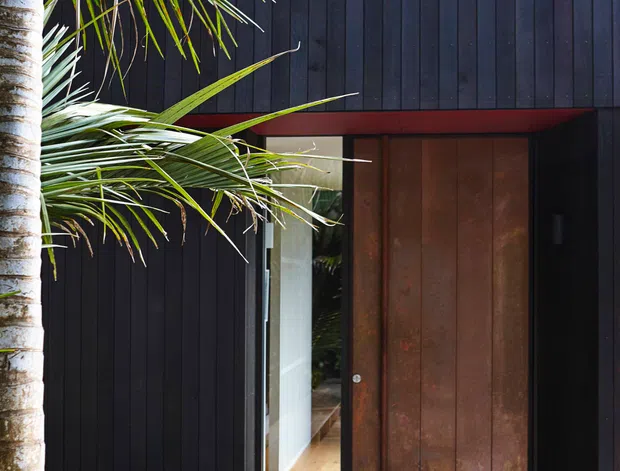 Exterior entrance to a contemporary dark-clad home, featuring vertical black timber cladding, a large rust-toned metal pivot door, glazed sidelighting, and timber decking with built-in bench seating. Tropical palm planting partially frames the facade, while the open doorway reveals a warm timber-floored interior.