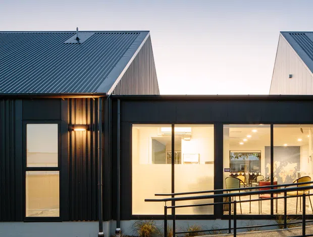 Exterior dusk view of a contemporary multi-volume residential building. Dark corrugated cladding forms the lower facade, with steep gabled roof volumes clad in ribbed metal and pale vertical siding. Large illuminated windows reveal a bright open-plan interior, while a black metal accessibility-style ramp and balustrade run across the foreground.