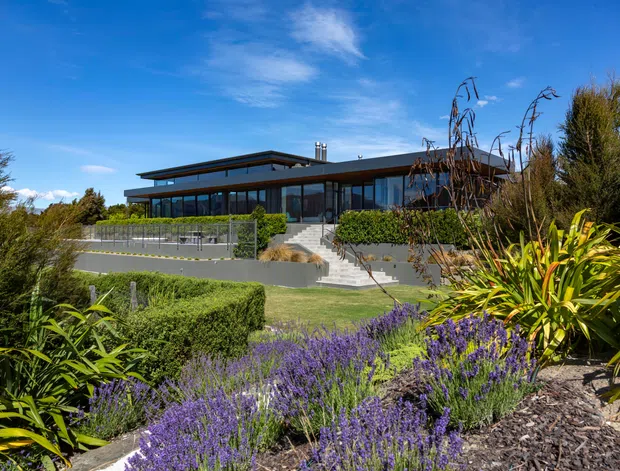 A low-profile contemporary building set within a landscaped garden, viewed from the front. The structure features broad dark-framed glazing, a flat projecting roofline, extensive outdoor terraces and steps, with clipped hedges, purple flowering plants and sculptural greenery in the foreground.