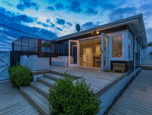 A dusk exterior view of a single-storey contemporary home with a timber deck and stepped timber boardwalk, open bi-fold doors, and a warmly lit interior visible through the glazing. The facade combines dark horizontal cladding with light trim, while a slatted privacy screen encloses an outdoor seating area and spa or plunge-pool enclosure. Shrubs and layered decking surround the raised terrace.