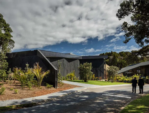 Exterior view of a contemporary dark-clad building complex set within landscaped grounds. The low-pitched angular rooflines, vertical dark wall cladding, exposed diagonal structural element, and broad paved driveway create a modern institutional or commercial architectural composition. Mature trees and ornamental planting frame the site, with a separate lighter-roofed building visible to the right.