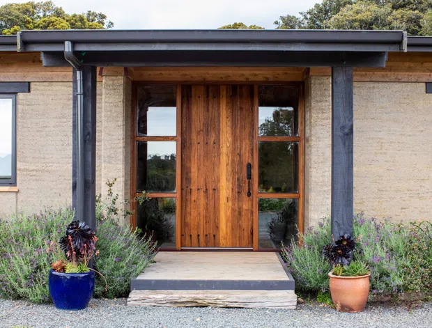 Front entrance of a low-profile single-storey dwelling with warm timber double doors, large glazed sidelights, textured light masonry walls, dark timber or metal structural posts, and a broad flat roof edge. Native-style landscaping and potted plants frame the entry path.