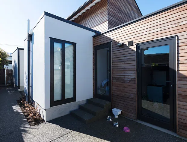 Exterior view of a compact contemporary residential building or extension, combining white rendered walls with warm horizontal timber cladding and dark-framed glazing. The irregular roof forms include a raised pitched section with a satellite dish, while a textured concrete-paved forecourt and narrow access path occupy the foreground.