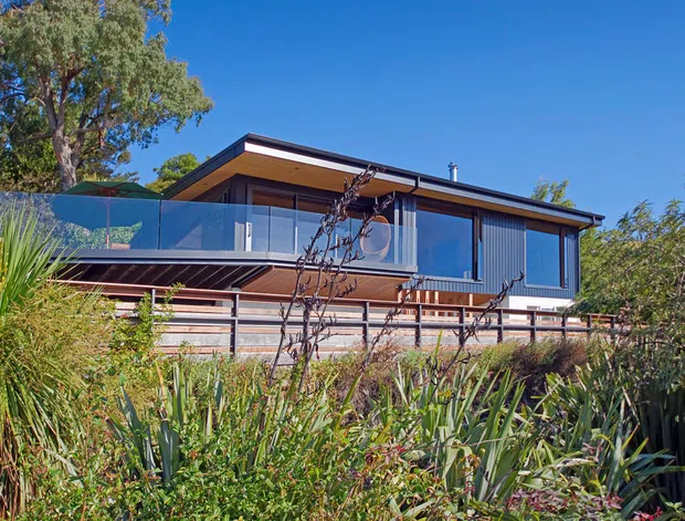 Exterior view of a contemporary elevated house set within dense landscaped planting. The building has a low-pitched roof, dark metal or timber-look cladding, extensive glazing, and a glass-balustraded upper terrace or balcony. Timber-toned horizontal screening and decking add warmth to the facade.