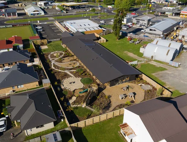 Aerial view of a contemporary low-rise building complex in a suburban mixed-use area. The central long dark-roofed building is surrounded by landscaped courtyards, winding paths, planted garden beds, seating areas, and fenced outdoor spaces. Detached homes and commercial or light-industrial buildings are visible around the site.