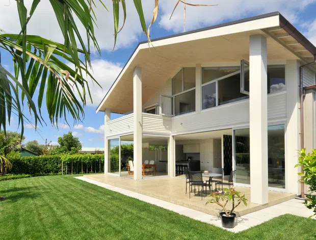 Exterior view of a contemporary two-storey residence with a broad low-pitched roof, deep eaves, white vertical cladding, slender structural columns, and extensive glazing. The upper level includes an open balcony or screened outdoor area, while the ground level opens onto a paved patio with outdoor dining furniture. A manicured lawn, hedging, and tropical planting surround the house.