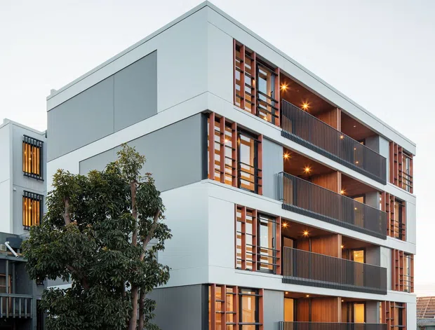 Street-level exterior photograph of a contemporary multi-storey apartment building. The building has a clean white and dark-grey facade, projecting balconies with dark metal balustrades, warm timber-lined balcony ceilings and vertical timber window screens, large glazed openings, and an integrated ground-level vehicle entry. Urban residential context with limited landscaping and neighbouring buildings visible.