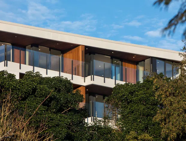 Exterior view of a contemporary multi-storey residence with a long, low flat roof, extensive floor-to-ceiling glazing, deep overhanging eaves, and warm timber-clad wall sections. Dark-framed glass balustrades and recessed outdoor areas are partially screened by mature landscaping.