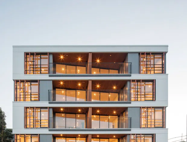 Front exterior view of a contemporary multi-storey apartment building with a regular stacked-balcony facade. The building has pale grey cladding, dark metal balcony balustrades, extensive glazing, warm illuminated interiors, and decorative vertical and horizontal metal screening around the windows. Street frontage, entry lobby, driveway access, and neighbouring urban buildings are visible.