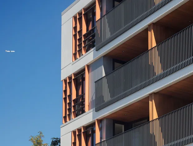 Exterior architectural photograph of a contemporary multi-storey apartment building against a clear blue sky. The facade features repeating recessed balconies with dark vertical metal balustrades, warm timber-lined soffits, pale grey structural framing, and orange-toned vertical sun-shading screens. Trees are visible at the lower edge.