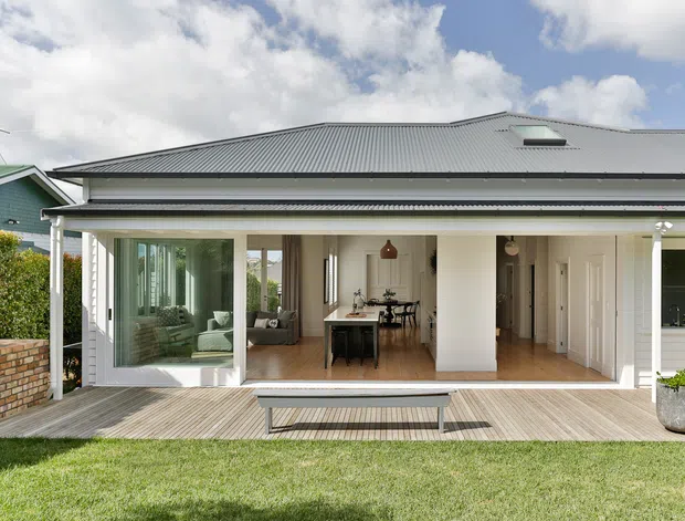 Rear exterior view of a light-coloured single-storey home with a broad covered veranda and open living connection to a timber deck. Large glazed sliding doors reveal a living area and dining space, while the corrugated metal pitched roof includes a skylight. A lawn and low brick boundary wall frame the outdoor setting.