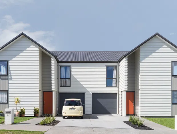 Front exterior view of a contemporary multi-unit residential building with three connected gabled volumes. The facade is clad in light horizontal weatherboards, with dark grey corrugated metal roofing, white-framed windows, and recessed red-orange entry doors. A central garage and driveway sit between the two taller end sections, creating a compact townhouse or apartment-style composition.