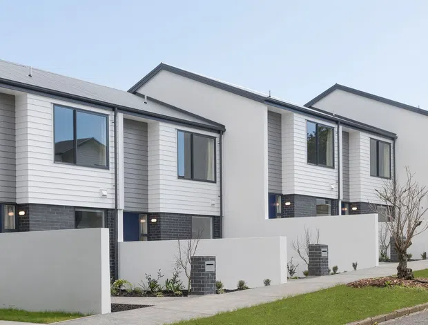 Exterior street view of a contemporary multi-unit townhouse development. The two-storey homes have repeated gabled roof forms, dark brick lower sections, light horizontal weatherboard-style upper cladding, dark-framed windows, recessed entry doors, and low white boundary walls along a landscaped footpath.