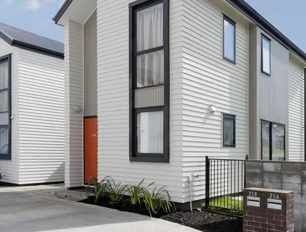 Exterior street view of a compact two-storey attached dwelling with white horizontal weatherboard cladding, a steep gabled roof, dark window and door frames, and a small landscaped front setback. The property includes a paved driveway, low brick letterbox, concrete block boundary wall, and black metal fencing.