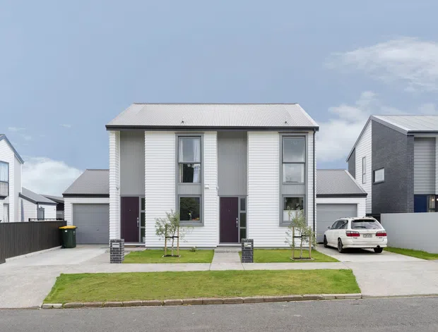 Street-facing view of a contemporary attached residential dwelling with a symmetrical two-storey facade. The home features light horizontal weatherboard cladding, dark brick or masonry accents, tall narrow windows, a pale corrugated pitched roof, small front lawns, concrete driveways, and neighbouring modern homes in a suburban development.