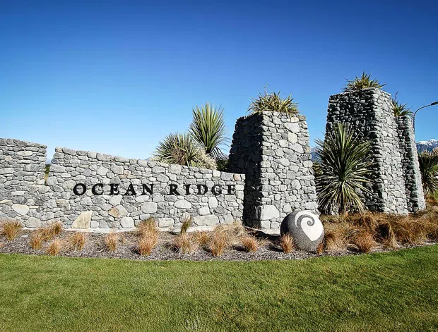 Exterior entrance sign for Ocean Ridge, formed from rough stacked grey stone walls and tall stone pillars, with sculptural stone elements and spiky ornamental plantings in a landscaped lawn. Clear blue sky and distant mountains are visible in the background.