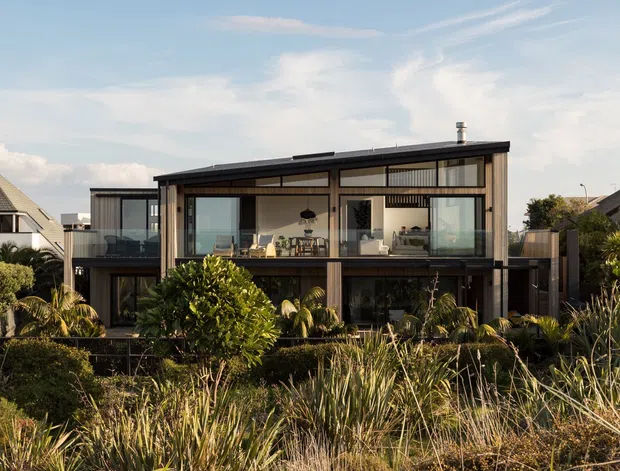 Exterior view of a contemporary two-storey house with a dark low-pitched roof, extensive floor-to-ceiling glazing, timber-toned vertical cladding, and projecting upper-level volumes. The home sits within landscaped coastal-looking planting, with a bright open sky and neighbouring houses visible around it.