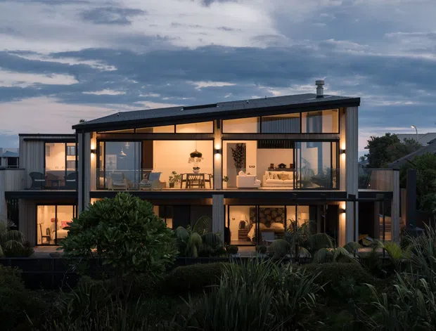 A contemporary two-storey residential house photographed at dusk, with a dark linear roof and structural framing, extensive floor-to-ceiling glazing, and warmly illuminated open-plan interiors. The home is surrounded by dense landscaped planting and neighbouring suburban homes.