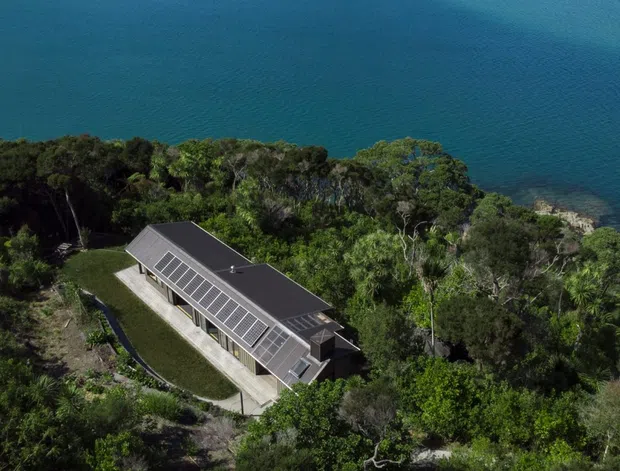 Aerial view of a long, contemporary single-storey building set among dense coastal vegetation beside deep blue water. The structure has a dark pitched roof with an extensive linear bank of roof glazing or skylights, light-toned exterior walls, and a narrow paved access path.