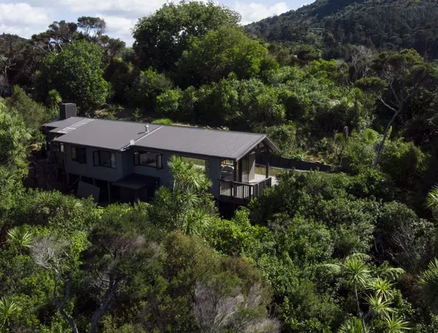 Aerial view of a dark-clad modern house nestled among dense native vegetation on a heavily forested, mountainous or sloped site. The home has a low-pitched roof, extensive glazing, and an elevated timber deck or balcony, with the surrounding landscape dominating the scene.