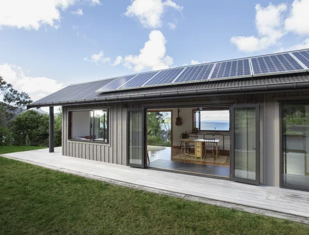 Exterior view of a single-storey contemporary house with a long pitched roof covered in solar panels, grey vertical cladding, extensive glazed sliding doors, and a pale timber deck. A dining area is visible through the open glazing, with a coastal or water view beyond. The house is set beside a lawn and trees under a bright sky.