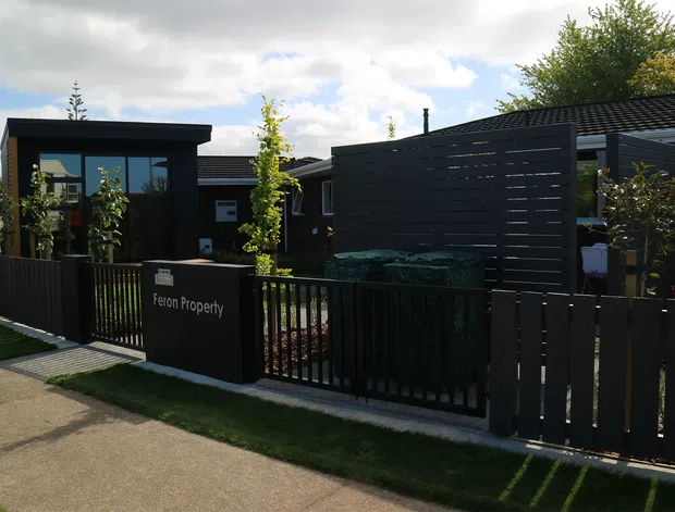 Street-facing view of a contemporary low-rise property with dark horizontal cladding, a flat-roofed entrance structure, slatted privacy screening, black fencing and landscaped planting. A sign identifies the property as Feron Property. The image is taken from the external walkway, with the building facade and front boundary dominating the frame.
