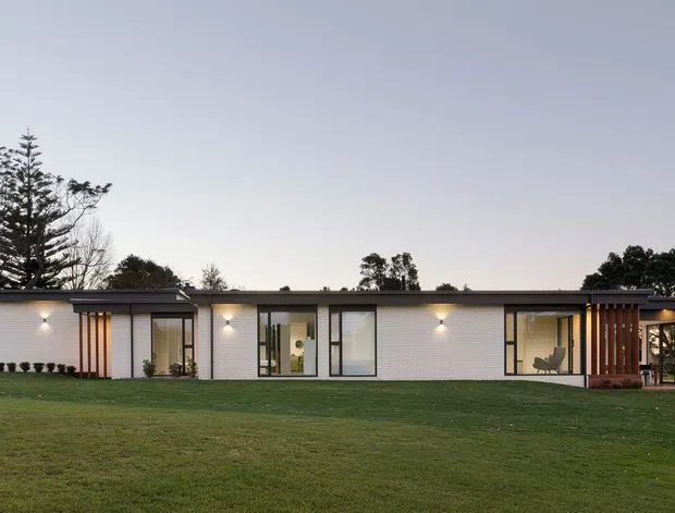 A low, single-storey modern house viewed from across a broad lawn at dusk. The long horizontal facade combines pale masonry or rendered wall panels with dark metal-framed glazing, a shallow flat roof, and warm exterior lighting. Timber-toned vertical slatted screens mark the entry and covered outdoor areas, with landscaped grounds and mature trees surrounding the building.
