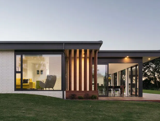 Single-storey contemporary residence viewed from the exterior at dusk, with a low flat roof, white brick masonry walls, dark-framed glazing, and a vertical timber screen at the junction between the enclosed living area and glazed outdoor-facing extension. Warm interior lighting reveals a furnished sitting area and dining space, while a broad lawn and mature trees surround the house.