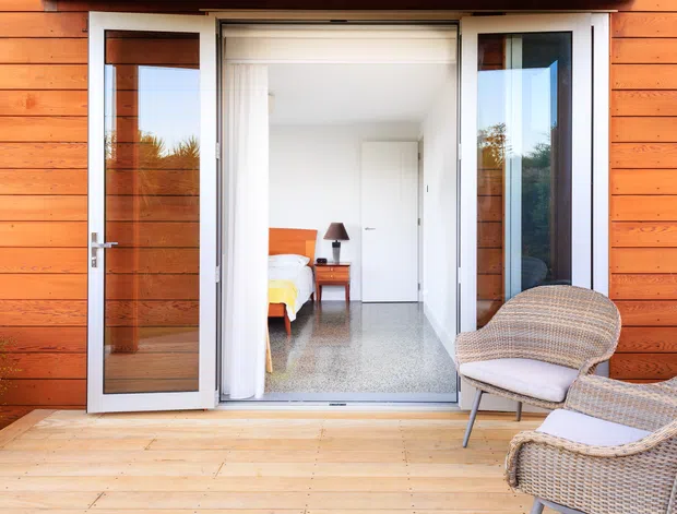 View from a timber deck through open glazed folding doors into a bright bedroom. The exterior facade is clad in warm horizontal timber boards, while the interior has white walls, a polished grey floor, and a partially visible bed with a bedside table and lamp. Wicker outdoor chairs occupy the foreground on the deck.