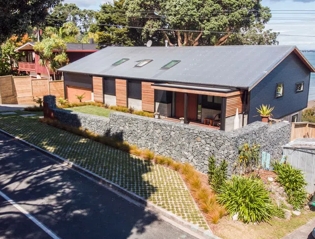 A contemporary single-storey coastal house viewed from the street, with dark grey metal cladding and roofing, warm timber-framed glazing, roof skylights, and a low gabion stone retaining wall. The house sits on a sloping site beside a sandy beach and visible water, with landscaped planting and permeable grassed paving at the frontage.