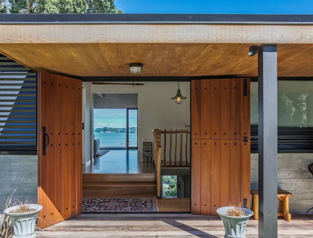 Exterior entrance of a contemporary coastal home, featuring wide timber pivot doors, a deep timber-lined entry canopy, dark horizontal cladding, exposed structural posts, and large glazed openings. The open doorway reveals a bright interior and a glimpse of water beyond.