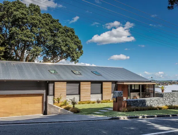 Street-facing exterior view of a contemporary single-storey home with a long pitched corrugated-metal roof, dark horizontal cladding, timber garage door and timber shutter panels, roof skylights, and a stone-clad front terrace wall. The home sits on a sloping suburban site with a visible waterfront or bay beyond.