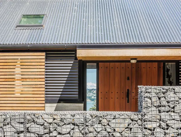 Exterior close-up of a contemporary house entrance with a corrugated metal roof, timber-clad front door and screen, dark horizontal facade elements, and gabion stone walls. Water and a distant shoreline are visible through the entry opening.