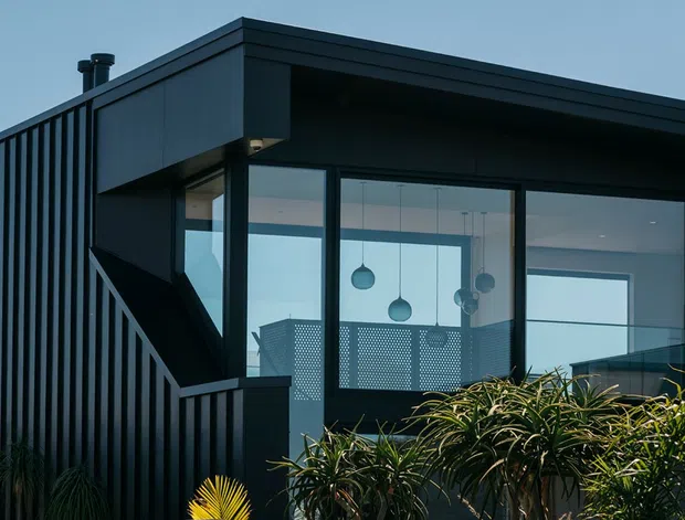 Exterior view of a contemporary, flat-roofed building with dark vertical cladding, broad black-framed glazing, and an external stair with matching balustrade. Pendant lights and interior spaces are visible through the windows, while dense ornamental planting softens the facade.