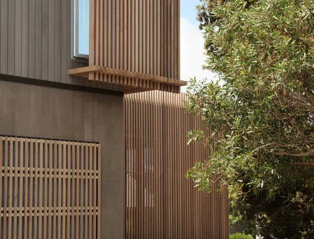 Exterior architectural view of a contemporary multi-level house featuring dark vertical cladding, extensive warm timber screening and a projecting screened window bay. A gabion stone retaining wall, timber slatted gate, concrete surfaces and established trees complete the composition.