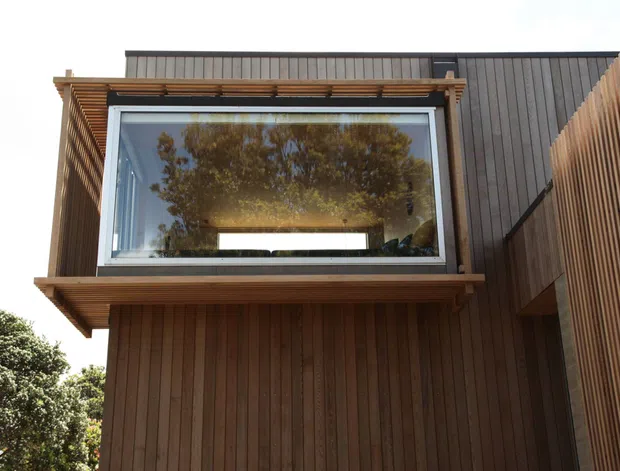 Exterior architectural view of a contemporary upper-level window projection or bay window. The facade is clad in vertical dark timber boards, with a projecting timber-framed glazed box and horizontal timber sun-shading slats. A dark standing-seam metal roof or upper wall plane is visible behind it, while trees are reflected in the glazing.