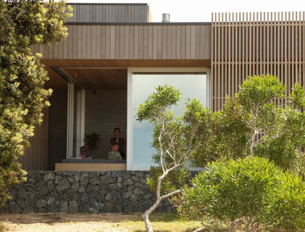 A contemporary residential facade viewed from outside, featuring vertical timber cladding and a prominent vertical timber screening element. A recessed entry or living area sits beneath a timber-lined soffit, with large glazing, a dark natural-stone retaining wall, and landscaped shrubs in the foreground. The low, rectilinear form and carefully screened openings create a private, modern exterior.
