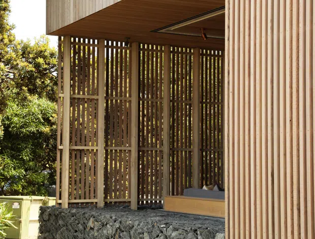 Exterior architectural view of a contemporary timber-clad structure with a deep covered outdoor area, vertical timber screening, exposed timber soffit, and a gabion-stone retaining wall. The open shelter includes a built-in bench and appears integrated into a landscaped site.