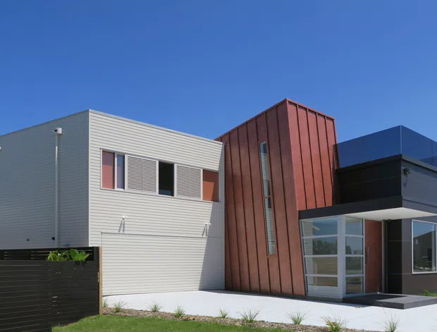 Exterior view of a contemporary multi-volume residence composed of contrasting geometric forms. The facade combines pale horizontal cladding, a tall rust-red vertical timber-clad volume with narrow clerestory-style glazing, and a dark charcoal box-like entry volume with large windows. A paved path, low dark boundary fence, and landscaped planting sit in front of the house under a clear blue sky.