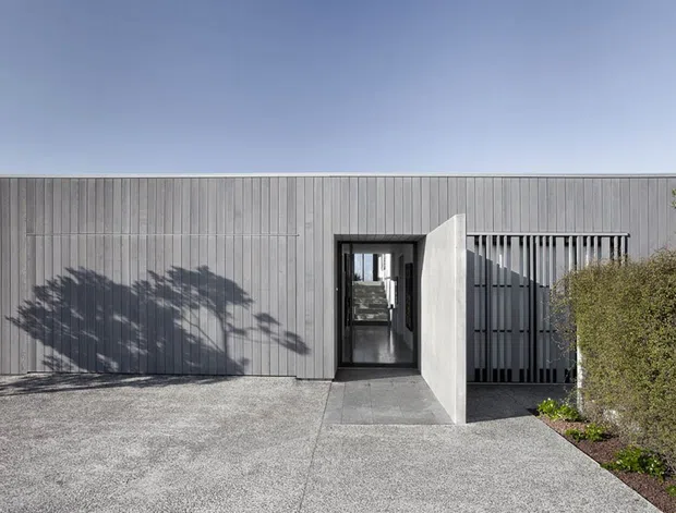A low, single-storey contemporary building viewed from the front, with a simple rectangular form, flat roofline, vertically ribbed light-grey cladding, dark-framed glazed entry, and a screened opening beside the entrance. A broad textured concrete driveway and sparse landscaping surround the facade.