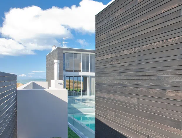 Exterior architectural view of a contemporary multi-level residence, focused on a tall dark timber-clad wall beside a narrow turquoise pool and glazed living space. White rendered walls, extensive glass, a rooftop edge, and a small grass courtyard are visible, with blue sky and distant landscape beyond.
