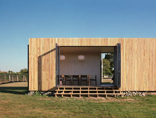 A compact single-storey rectangular building set in an open rural landscape, clad in vertical light timber boards. A large central opening has dark-framed folding or sliding doors, revealing a simple dining area with a table, chairs and pendant lights. A small timber deck and steps sit in front of the opening, with exposed stone edging at the base. The image has a clean, contemporary yet rustic character under a clear blue sky.