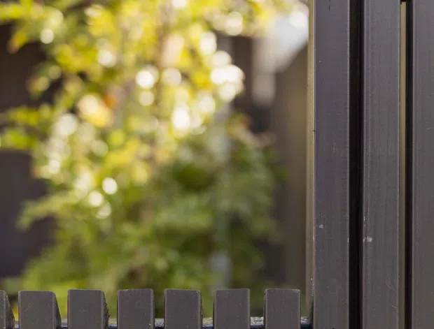 Close-up exterior view of a dark grey vertical slat fence or screen, with a matching taller privacy panel at right. Softly blurred greenery and a building facade appear in the background, creating a shallow-depth-of-field architectural product-style image.
