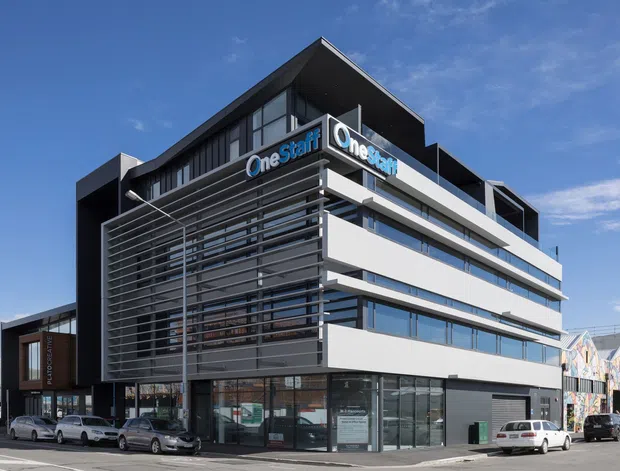 A contemporary multi-storey commercial office building on a street corner, featuring a dark and white facade, extensive horizontal glazing, prominent external metal sun-shading screens, and rooftop signage. Ground-level storefronts and parked vehicles line the surrounding urban streetscape.