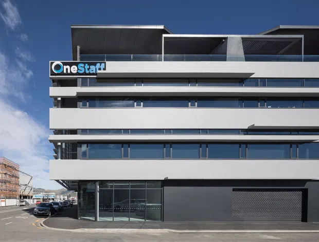 Street-facing view of a contemporary multi-storey commercial building with a dark charcoal and white facade, extensive horizontal bands, glazed office-level windows, rooftop terrace structures, and a ground-floor glazed entrance. A OneStaff sign is mounted near the upper facade, with neighbouring urban construction visible at the side.