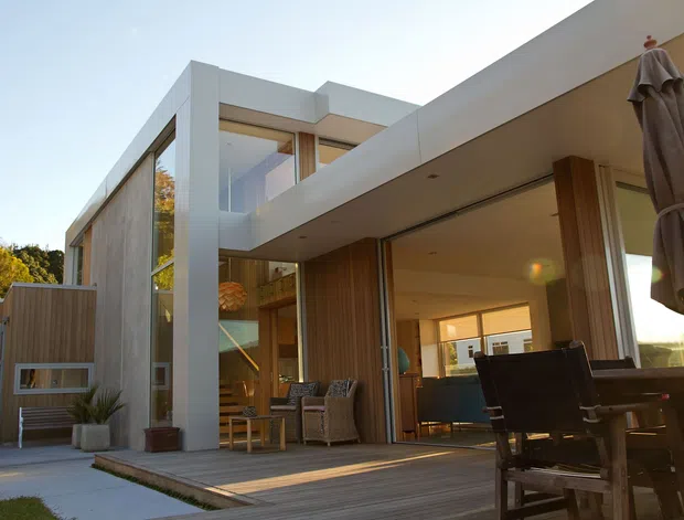 Contemporary two-storey residential home viewed from an outdoor entertaining area, with strong white geometric volumes, extensive glazing, timber-clad walls, a timber deck, and an open-plan interior visible through large sliding doors. Outdoor dining furniture and a closed umbrella occupy the foreground.