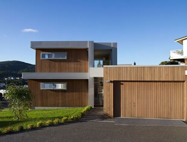 Exterior view of a modern two-storey residence with a flat roof, warm vertical timber cladding, crisp concrete-coloured projecting slabs, narrow horizontal windows, and an integrated timber-clad garage. The home sits on a suburban sloping site with a paved driveway and landscaped lawn.