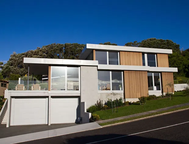A contemporary two-storey residence on a sloping suburban site, featuring a clean white and grey rendered facade, extensive glazing, warm vertical timber screening, flat roof planes, and an integrated double garage. The home is surrounded by mature trees and a neat lawn, with a street and driveway in the foreground.